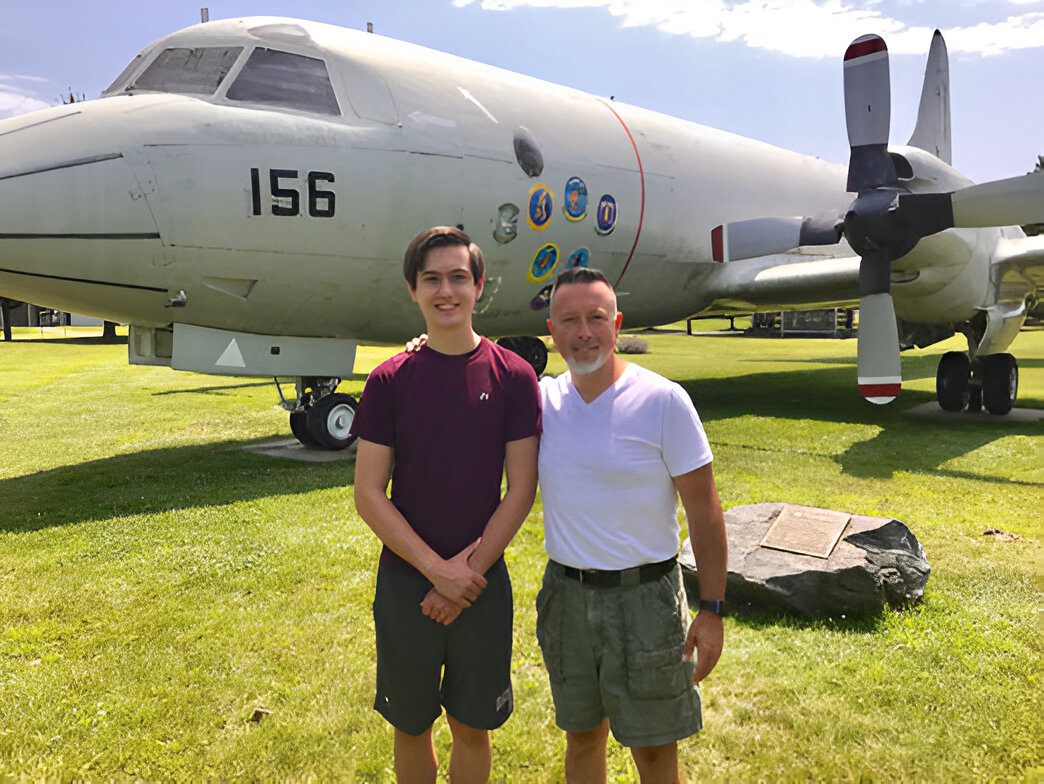 Two people stand on grass in front of a large aircraft with markings and the number 156. They are smiling and posing for the photo.