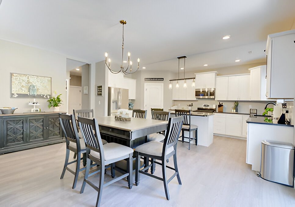 Modern kitchen and dining area with a wooden table and chairs, white cabinets, stainless steel appliances, and a chandelier. Light wood flooring and neutral walls enhance the bright space.